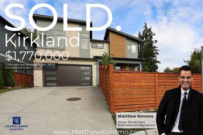 Modern House in Kirkland WA sold by Matthew Konsmo Realtor. A modern two-story home with a sleek, contemporary design, featuring a mix of materials such as light stone, wood paneling, and gray siding. The house has a large, dark brown garage door with frosted glass panels and an expansive concrete driveway. A wooden horizontal slat fence surrounds the property, providing privacy and complementing the home's modern aesthetic. The front yard includes a small patch of grass with young trees along the sidewalk. The house is situated in a suburban neighborhood, with neighboring homes visible, and the sky above is partly cloudy.