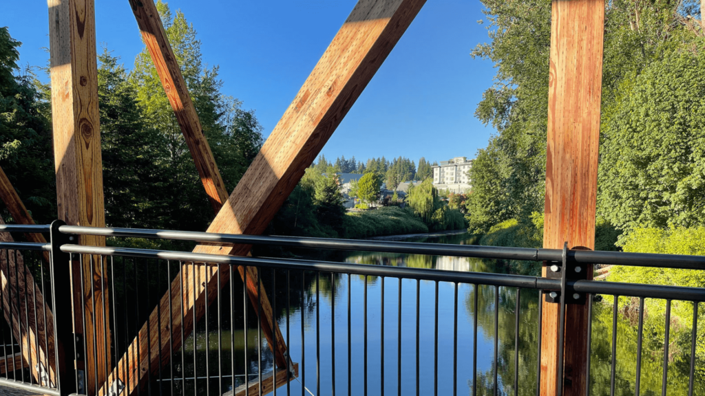 An arched wooden pedestrian bridge spanning the Sammamish River at the Park at Bothell Landing, connecting downtown Bothell to the regional trail system.