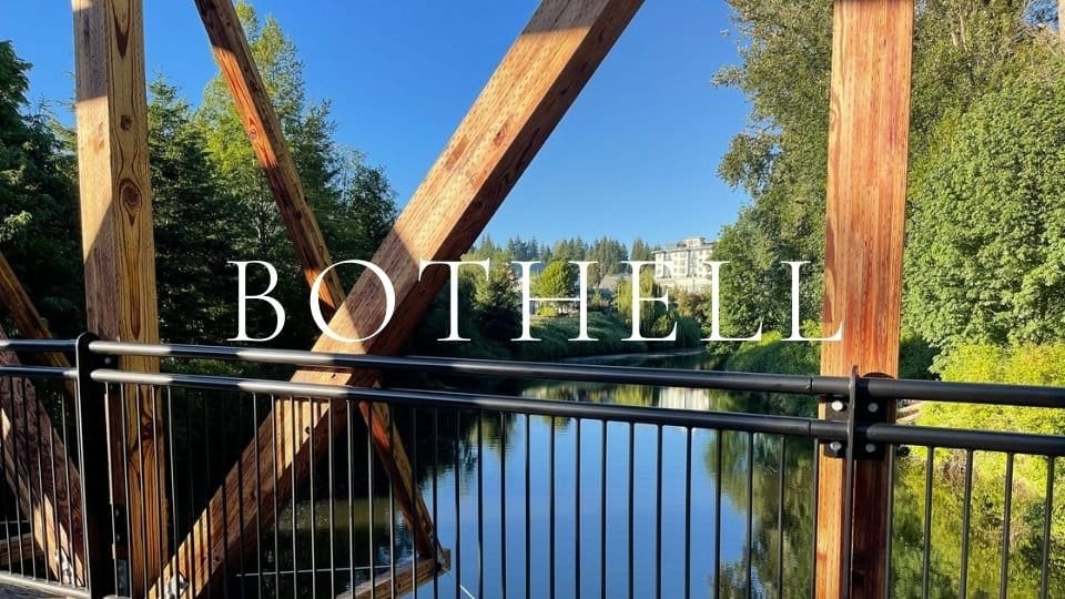 Wood truss pedestrian bridge in downtown Bothell crossing the Sammamish River near the Burke-Gilman Trail with greenery and condos in the distance.
