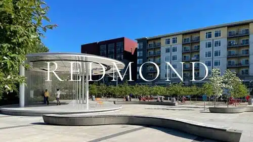 Downtown Redmond Park public plaza in Redmond Washington, featuring the iconic curved "REDMOND" branded wall, modern mixed-use apartment towers behind, blooming dogwoods, and stepped seating — Redmond WA real estate city guide imagery courtesy of Matthew Konsmo, Coldwell Banker Danforth.