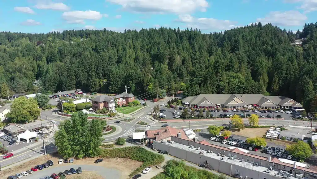 Wide aerial drone view of the Hollywood Hill Schoolhouse and surrounding winery district in Woodinville Washington, with shop blocks, the iconic roundabout intersection, and densely forested foothills beyond — Woodinville WA real estate city guide imagery courtesy of Matthew Konsmo, Coldwell Banker Danforth.