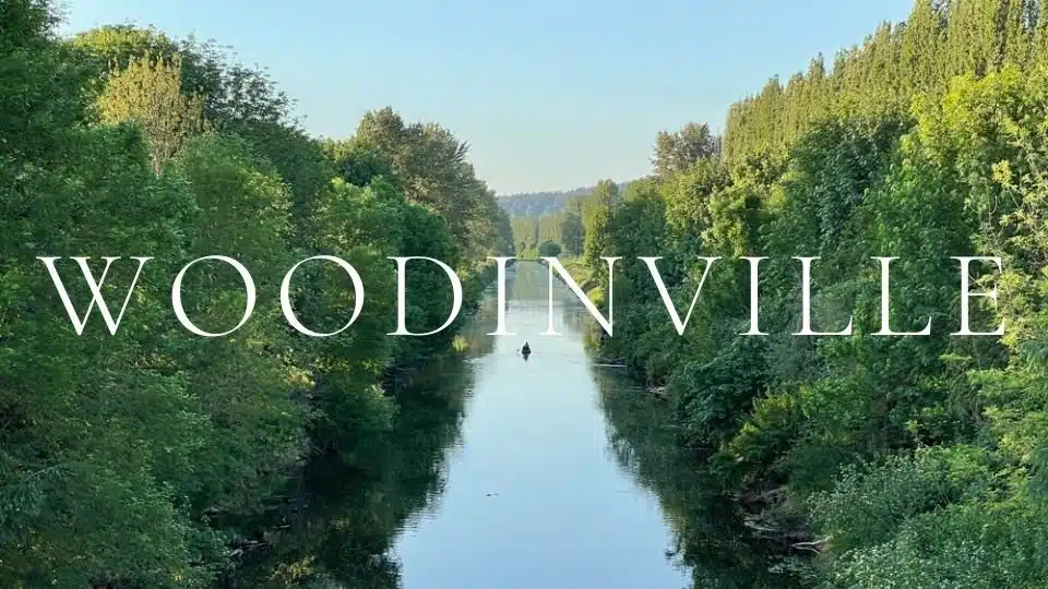 A lone kayaker paddles down the calm Sammamish River through a lush green valley in Woodinville, WA, with reflective water and dense trees lining the banks.