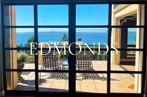 Edmonds, WA waterfront home interior view through grid-pattern French doors to a tiled terrace with patio seating and panoramic Puget Sound and Olympic Mountain views.