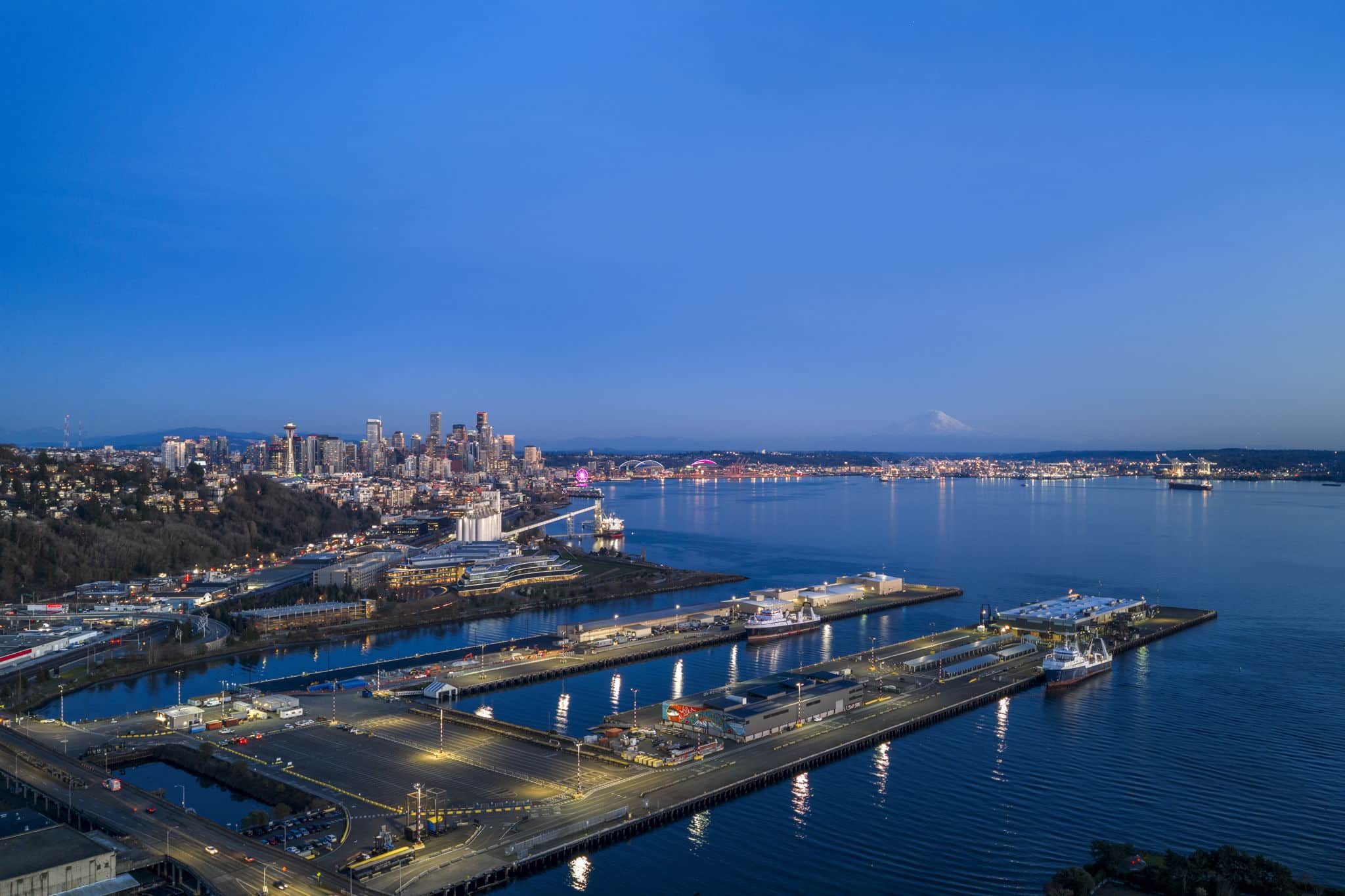 Aerial twilight view from Magnolia Seattle luxury home at 1740 Magnolia Way W showing panoramic Elliott Bay waterfront, downtown Seattle skyline with Space Needle, Seattle Great Wheel, Port of Seattle piers, and snow-capped Mount Rainier in the distance — listed by Matthew Konsmo Coldwell Banker Danforth Seattle real estate agent
