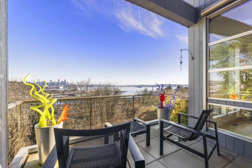 Covered outdoor balcony at Magnolia Seattle luxury home 1740 Magnolia Way W featuring colorful art glass sculptures, black mesh lounge chairs, cable railing, and unobstructed views of Space Needle, downtown Seattle skyline, and Elliott Bay on a sunny day — listed by Matthew Konsmo Coldwell Banker Danforth Seattle real estate agent