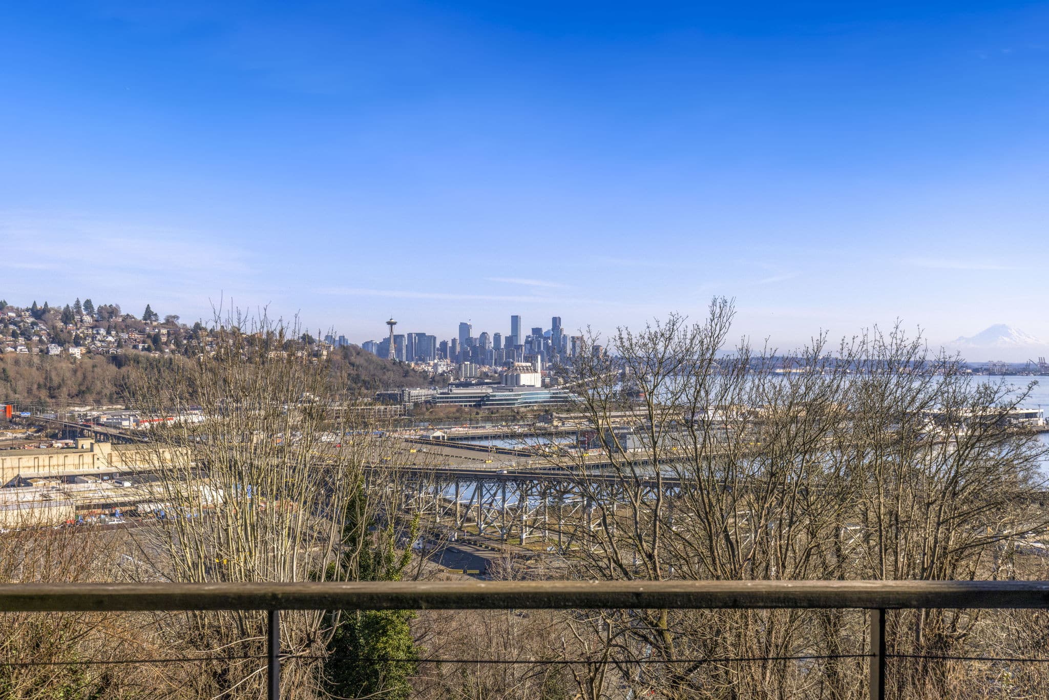 Outdoor deck view from Magnolia Seattle luxury home at 1740 Magnolia Way W featuring sweeping 180-degree panorama of Space Needle, downtown Seattle skyline, Elliott Bay waterfront, Magnolia Bridge, and snow-capped Mount Rainier on a clear blue sky day — listed by Matthew Konsmo Coldwell Banker Danforth Seattle real estate agent