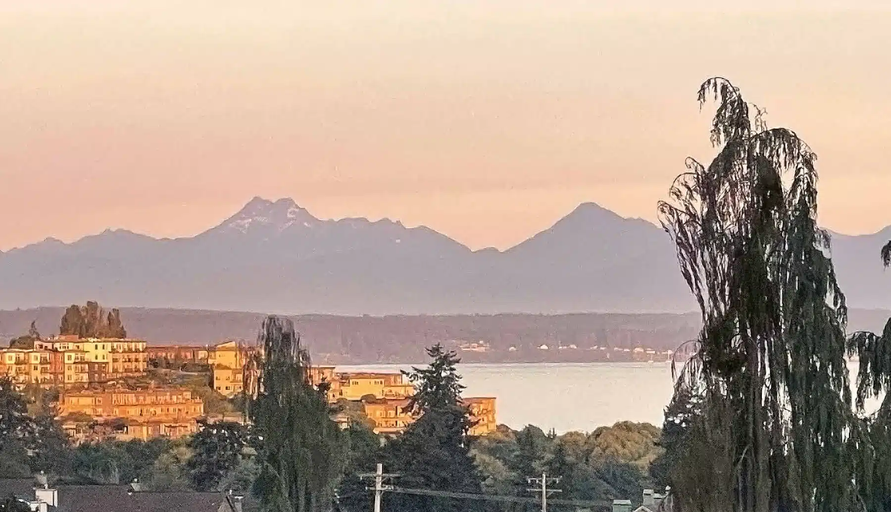 Elevated view of downtown Edmonds, Washington at sunset, overlooking the Puget Sound with the Olympic Mountains visible in the background under a warm, soft sky.