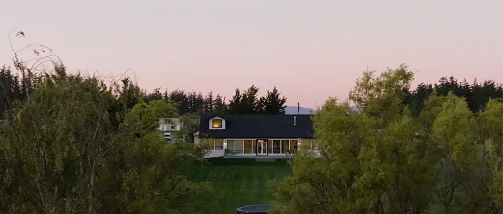 A modern single-family home with a dark charcoal roof and white siding, nestled on a manicured 5-acre green lawn on Camano Island. Framed by leafy green trees under a soft, twilight sky at sunset, the house features lit windows and large glass doors looking out over the property.