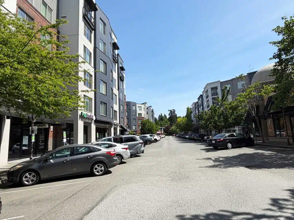 Living in South Juanita, Kirkland: 2026 Neighborhood & Real Estate Guide 3 Wide street view of mixed-use condominium buildings, retail storefronts, and angled parking at Juanita Village in Kirkland WA.