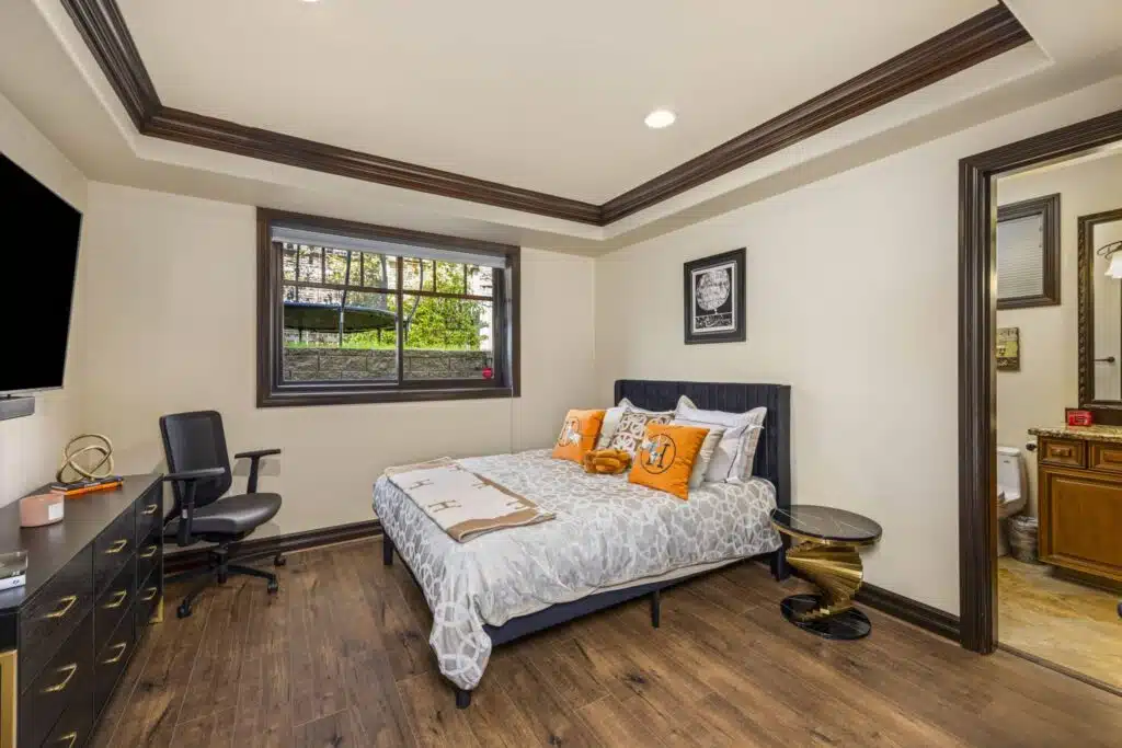 Spacious lower-level bedroom featuring tray ceiling with rich wood detailing, large picture window, and access to en-suite bathroom.