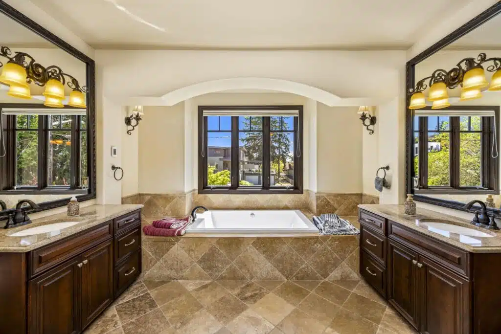 Spa-inspired Primary bath with deep soaking tub set beneath picture windows, framed by dual vanities with rich wood cabinetry.