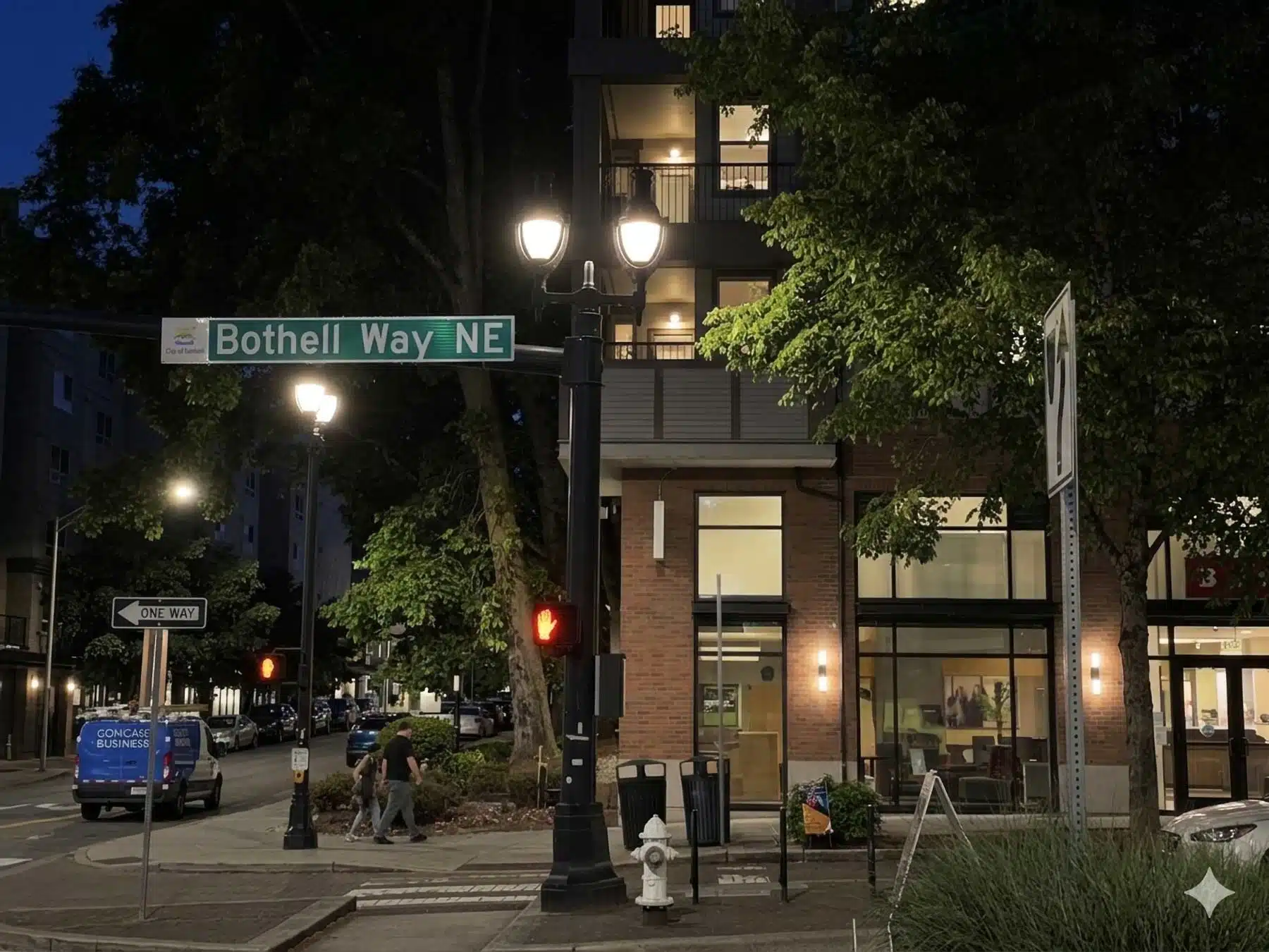 A nighttime street scene at a crosswalk in downtown Bothell, Washington, showing a prominent green street sign for Bothell Way NE attached to an illuminated ornate lamppost, with pedestrian traffic and a modern mixed-use brick apartment building in the background.