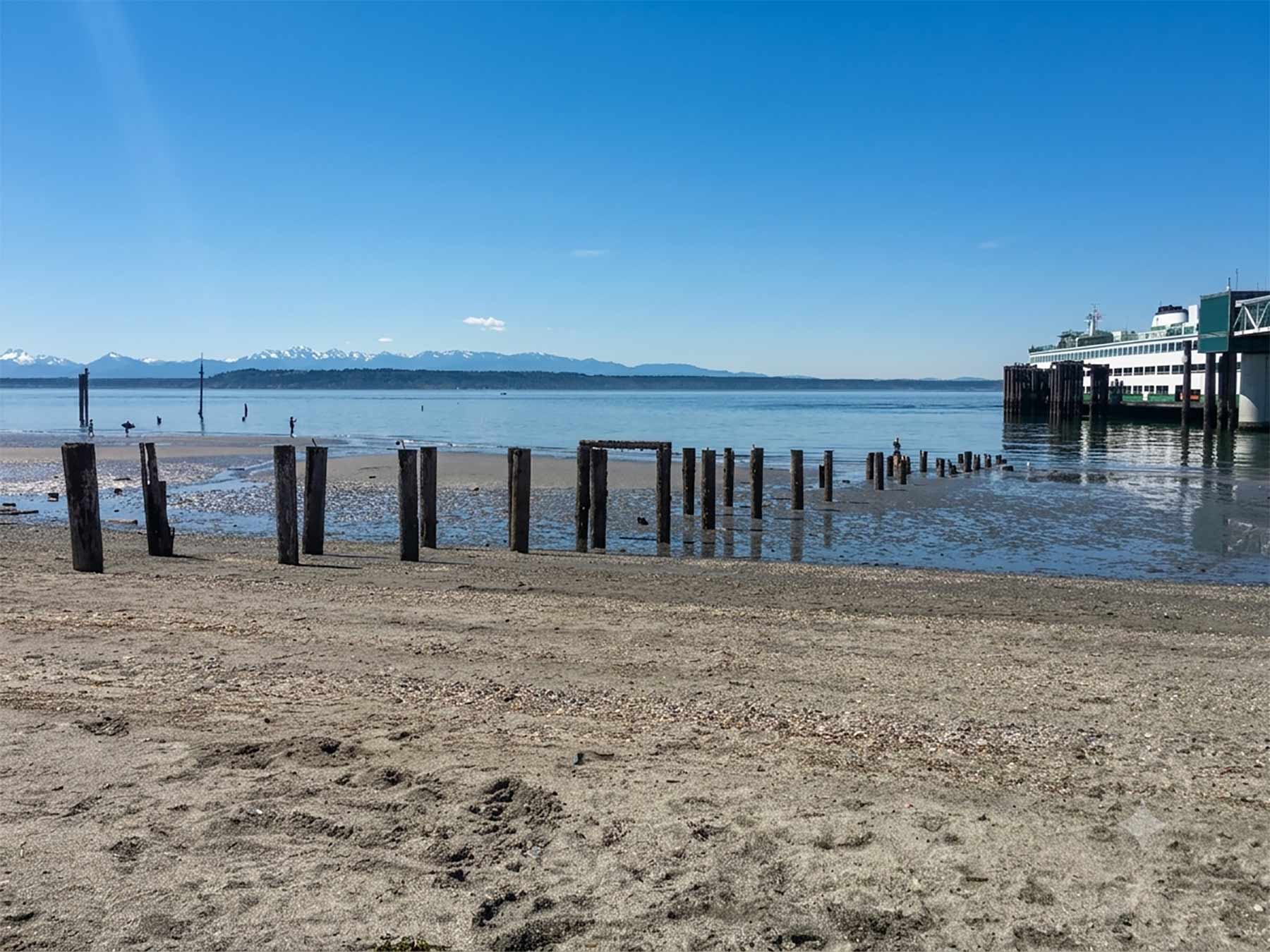 Living in Edmonds, WA - Your 2026 Neighborhood & Real Estate Guide 7 View of the sandy beach at Downtown Edmonds Waterfront Park, featuring weathered wooden pilings in the water, the Edmonds-Kingston ferry docked at the terminal, and the Olympic Mountains under a clear blue sky.