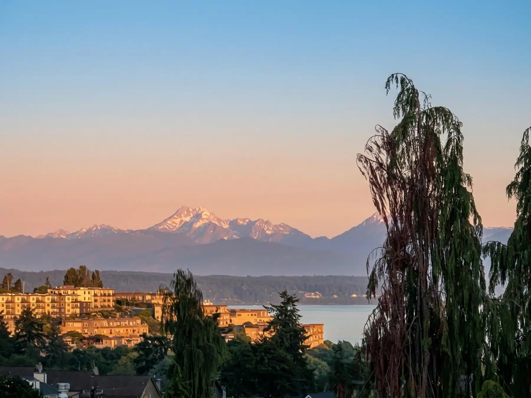 An elevated, scenic landscape photograph looking across the hillside residential communities of Edwards Point in Edmonds, Washington, toward the calm waters of Puget Sound and the snow-dusted Olympic Mountains illuminated by a warm sunset glow.