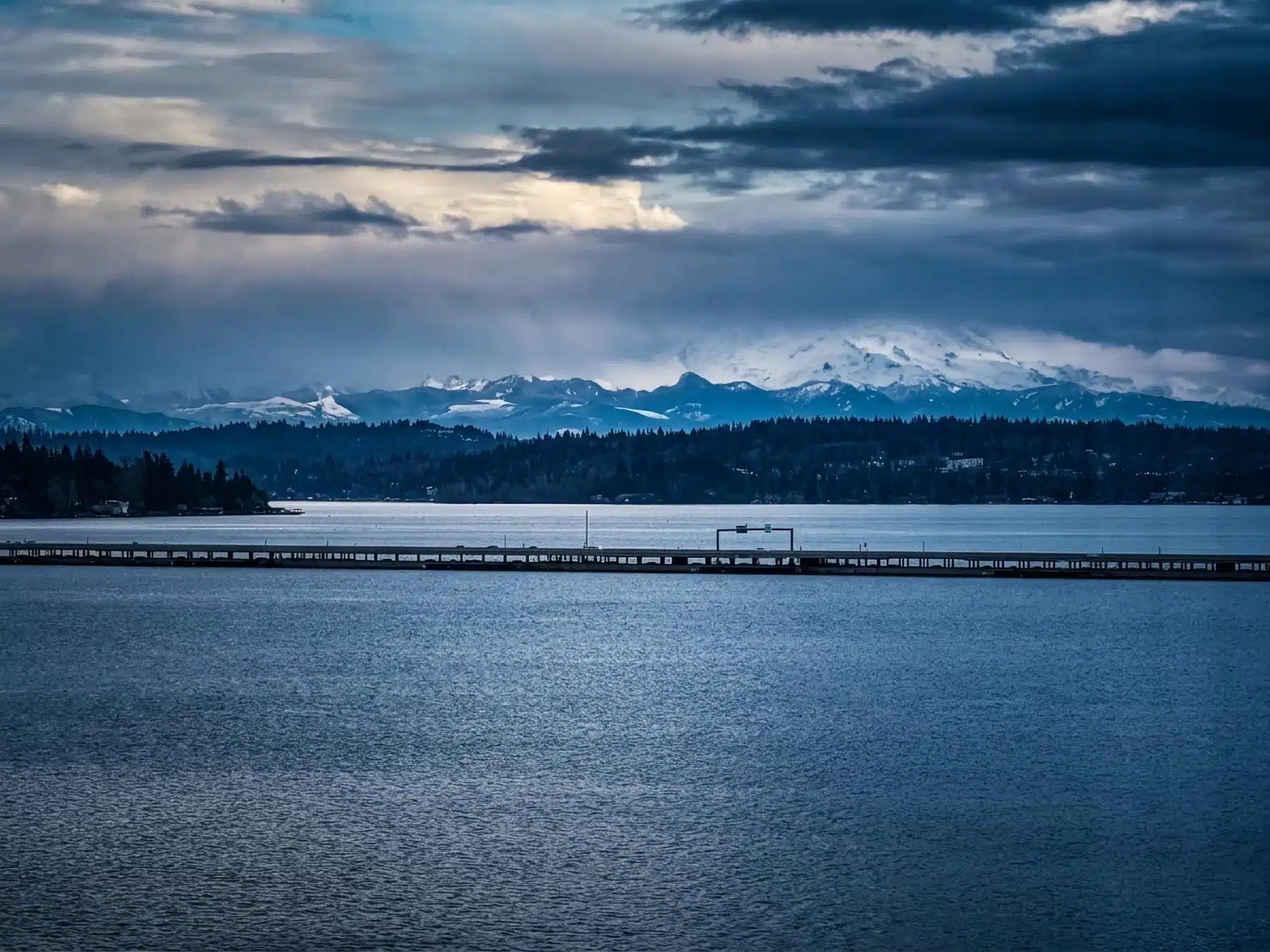 A wide landscape photograph taken from Seattle's Laurelhurst neighborhood, looking across the blue waters of Lake Washington and the SR 520 floating bridge toward a tree-lined shoreline, with a snow-covered Mount Rainier rising under dramatic, moody storm clouds.