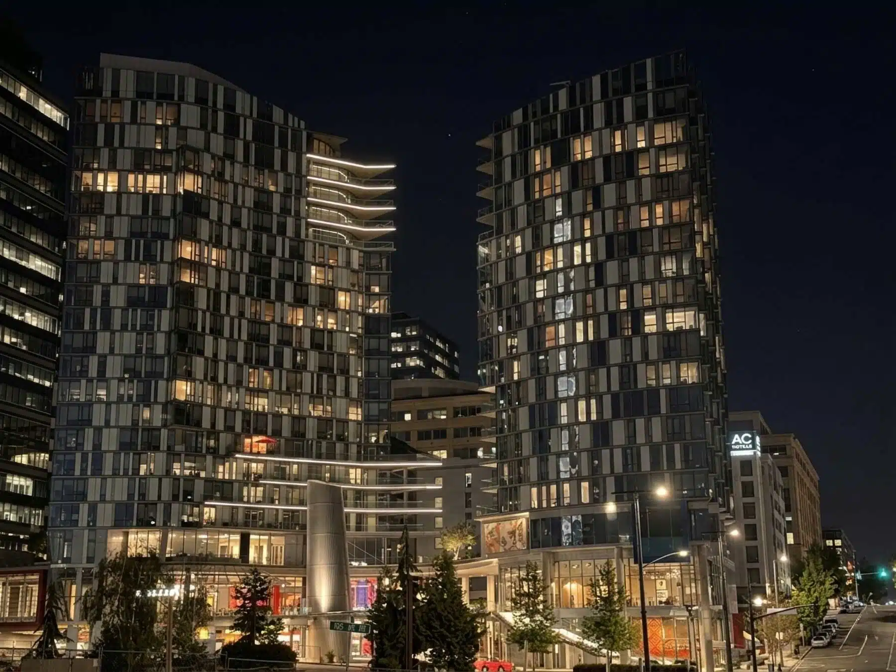 Living in Downtown, Bellevue: 2026 Neighborhood & Real Estate Guide 1 A wide-angle nighttime photograph of downtown Bellevue, Washington, featuring the illuminated, modern architectural facade of the dual high-rise towers at the Bellevue Avenue and NE 10th Street intersection, with streetlights and an AC Hotel sign visible.