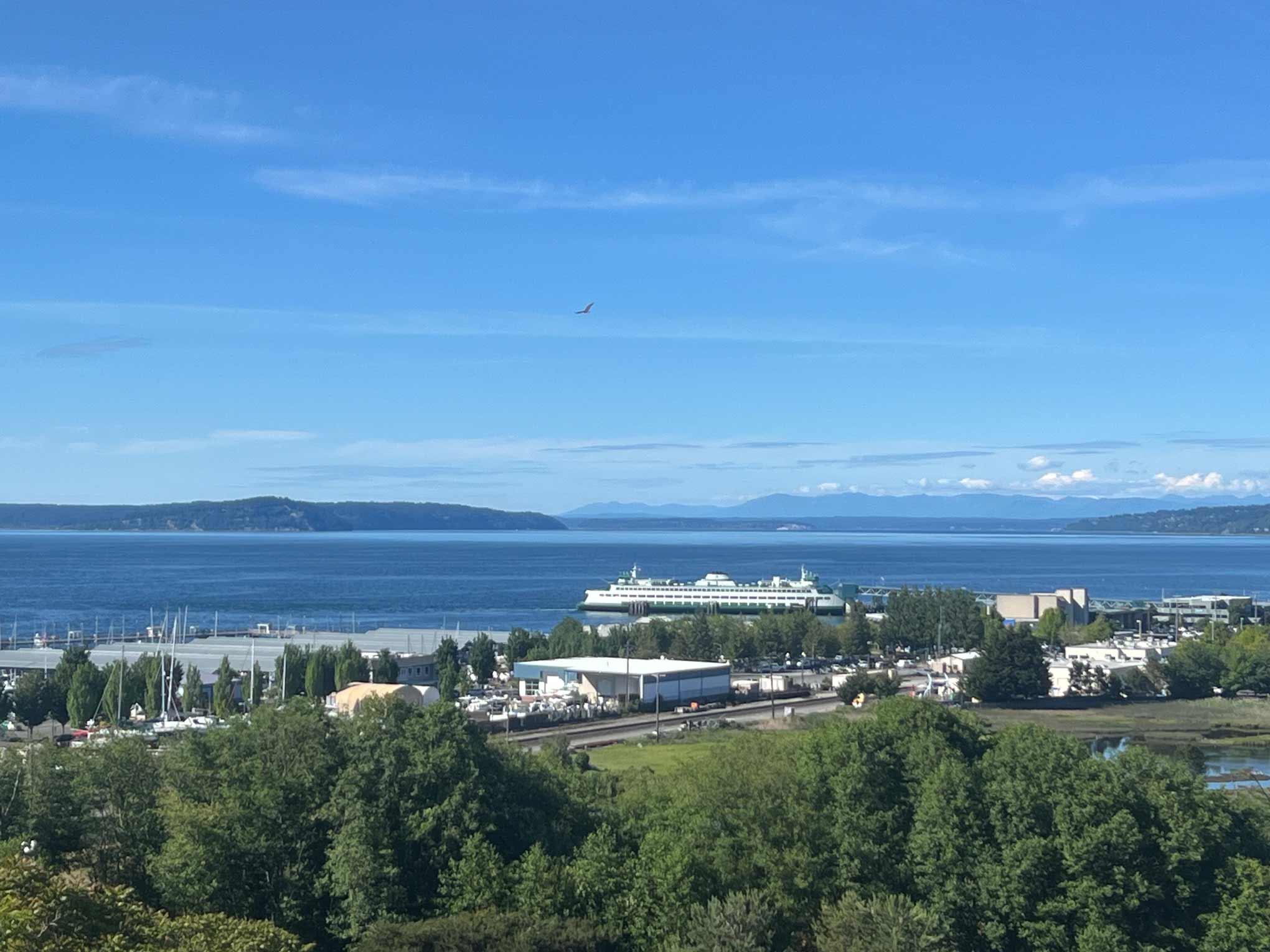 Living in Edmonds, WA - Your 2026 Neighborhood & Real Estate Guide 4 Daytime view of the Edmonds waterfront with a green-and-white Washington State ferry docked at the terminal, the Port of Edmonds marina to the left, and the Olympic Mountains rising across Puget Sound under a clear blue sky.