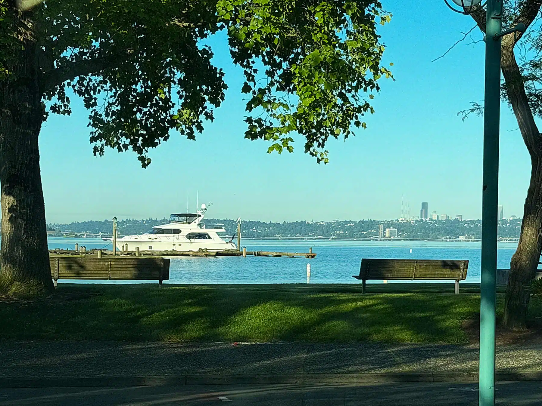 View of a luxury yacht docked at Marina Park in Kirkland, Washington, framed by park trees and benches with the Seattle skyline visible across Lake Washington.