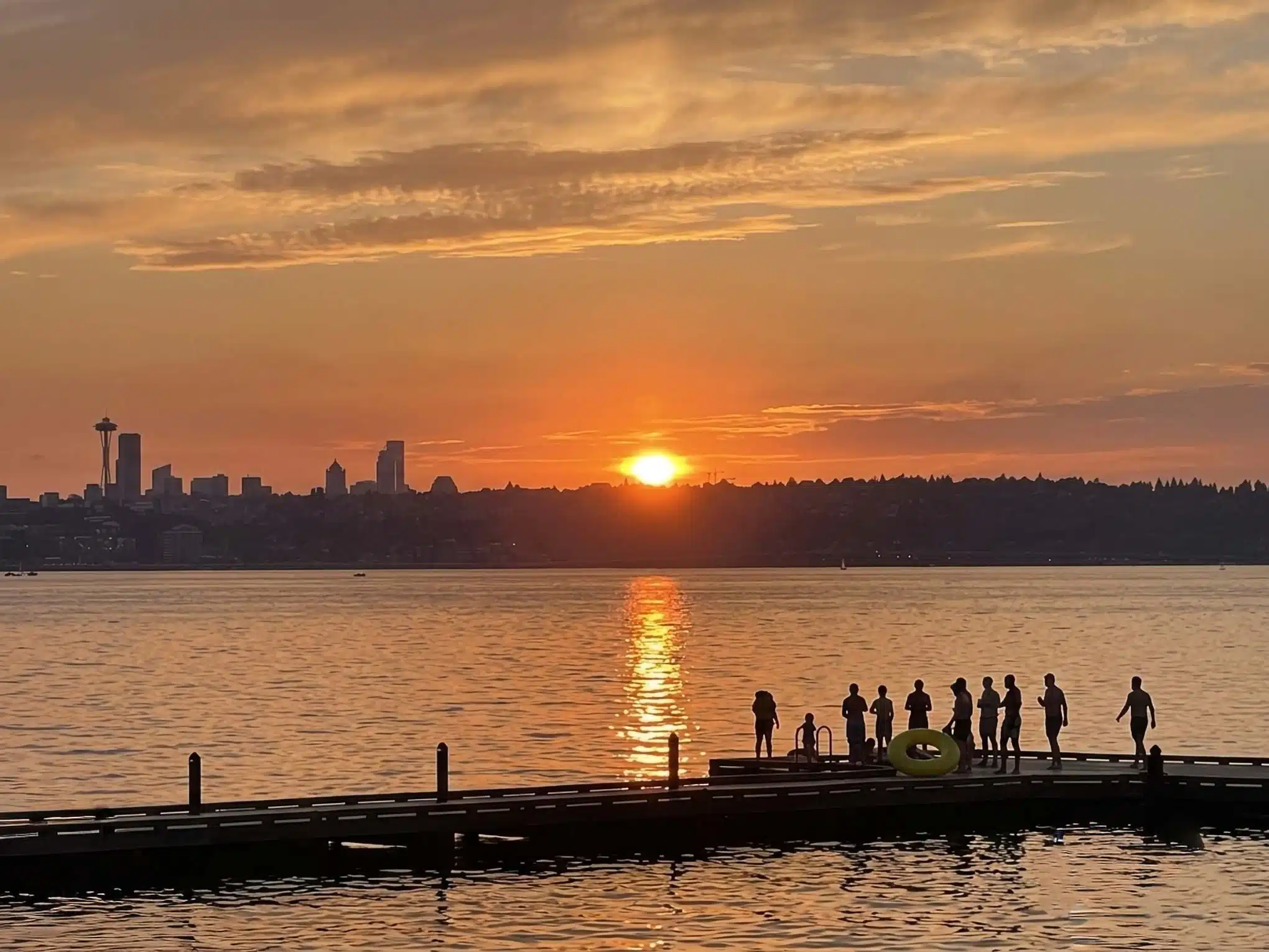 Sunset over Lake Washington from the dock at Waverly Beach Park in Kirkland, with the Seattle skyline and Space Needle silhouetted across the water as swimmers gather on the dock.