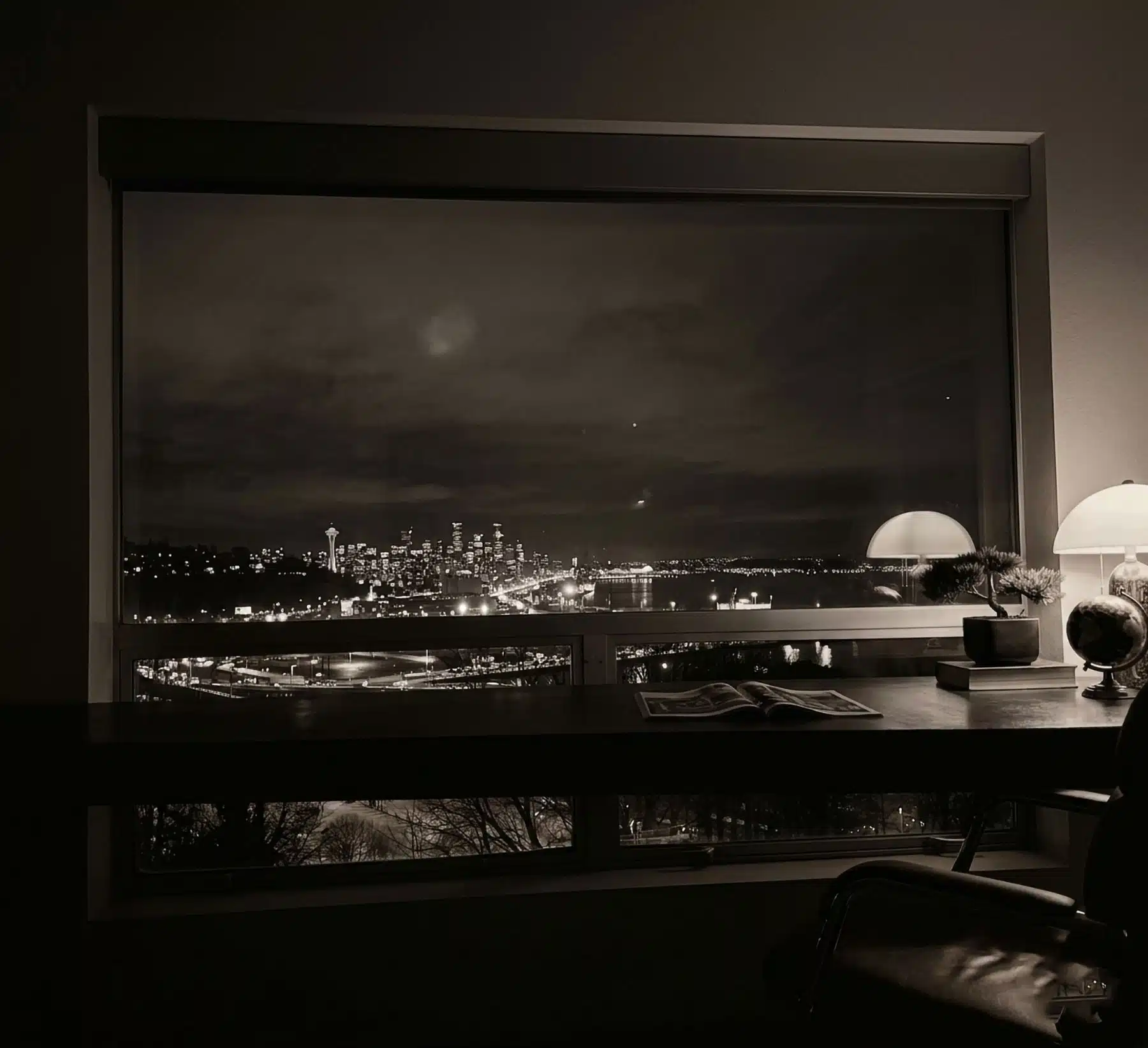 A black and white interior photograph looking through a large picture window from a home office desk, showcasing a dramatic nighttime view of the illuminated downtown Seattle skyline, the Space Needle, and Elliott Bay from the Magnolia neighborhood.